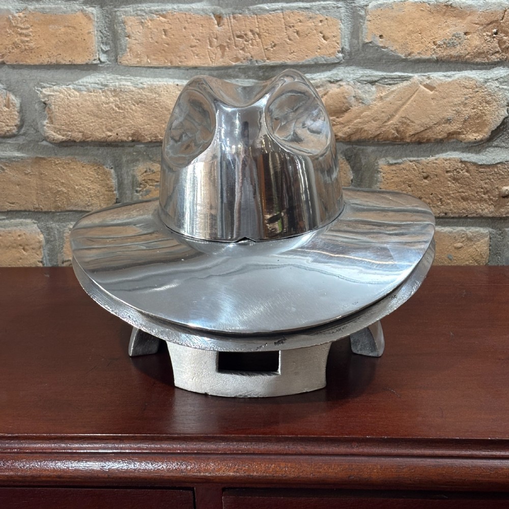A polished silver aluminium milliners hat block mould shaped like a fedora resting on a wooden surface in front of a brick wall. View II