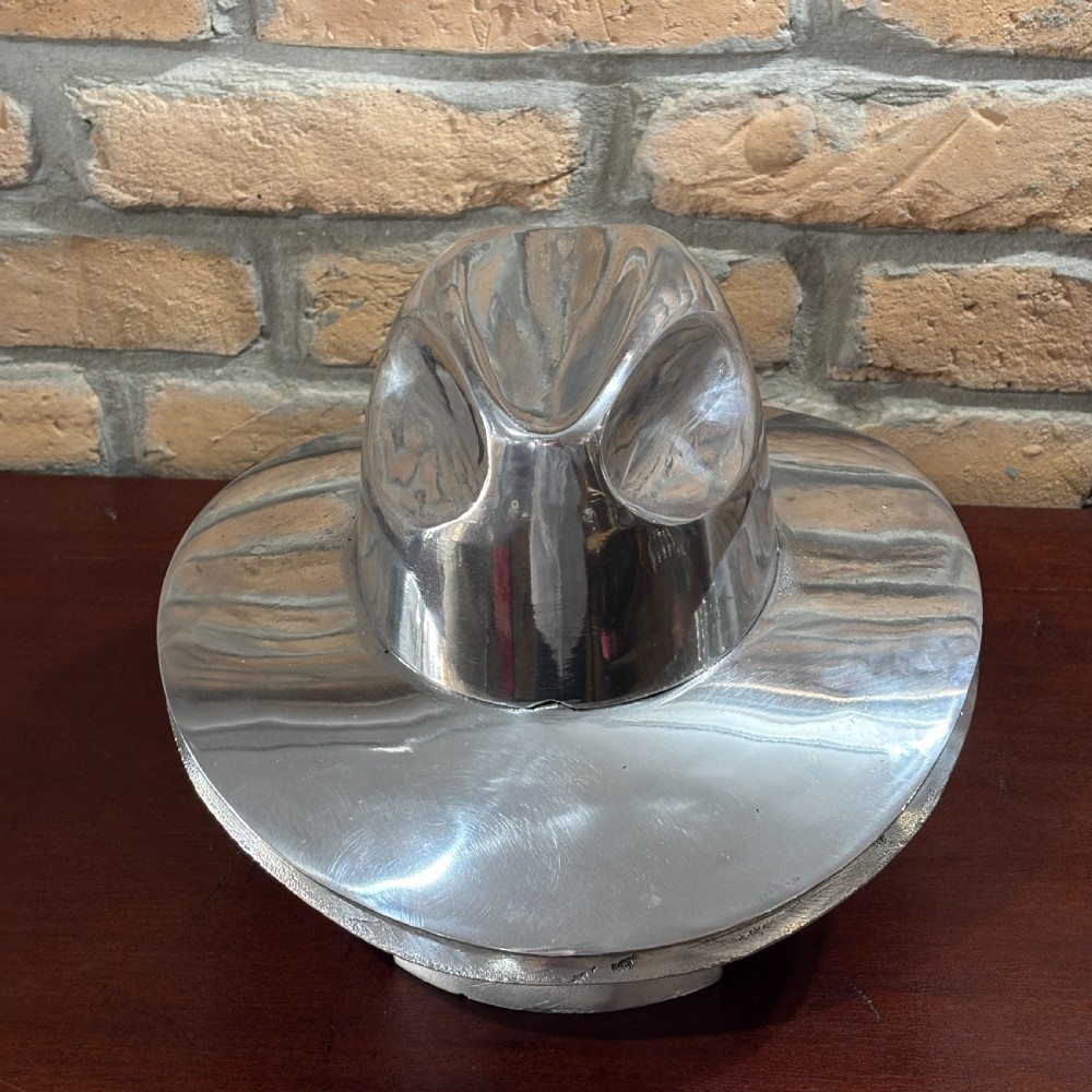 A polished silver aluminium milliners hat block mould shaped like a fedora resting on a wooden surface in front of a brick wall. View III