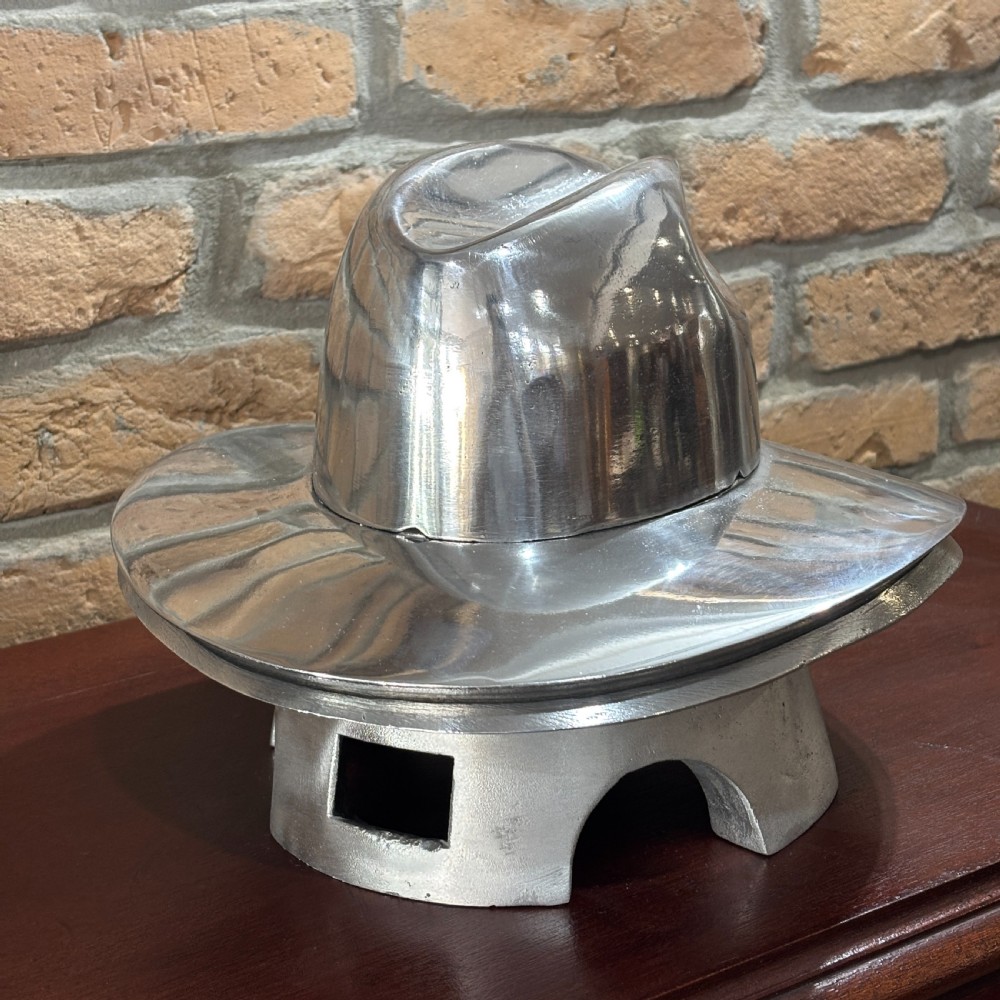 A polished silver aluminium milliners hat block mould shaped like a fedora resting on a wooden surface in front of a brick wall. View IV