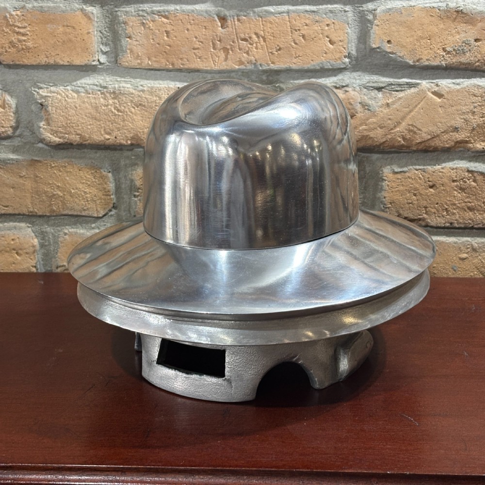 A polished silver aluminium milliners hat block mould shaped like a fedora hat resting on a dark timber surface.
