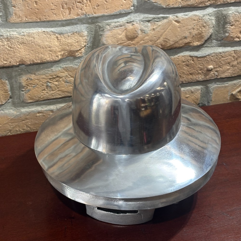 A polished silver aluminium milliners hat block mould shaped like a fedora hat resting on a dark timber surface. View II