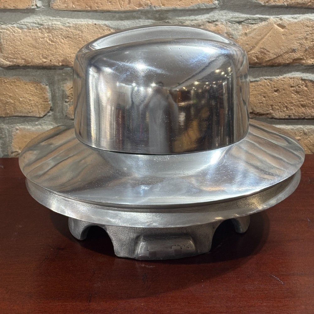 A polished silver aluminium milliners hat block mould shaped like a fedora hat resting on a dark timber surface. View III