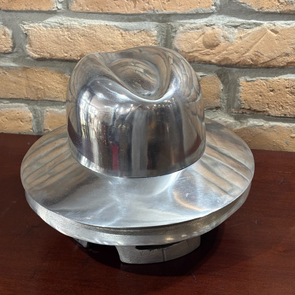 A polished silver aluminium milliners hat block mould shaped like a fedora hat resting on a dark timber surface. View IV