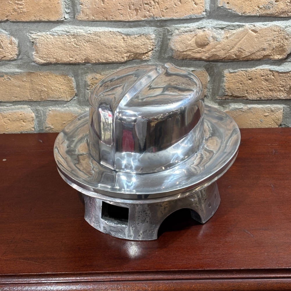 A polished cast aluminium millinery hat block mould forming a fedora shape on a contoured pedestal base.