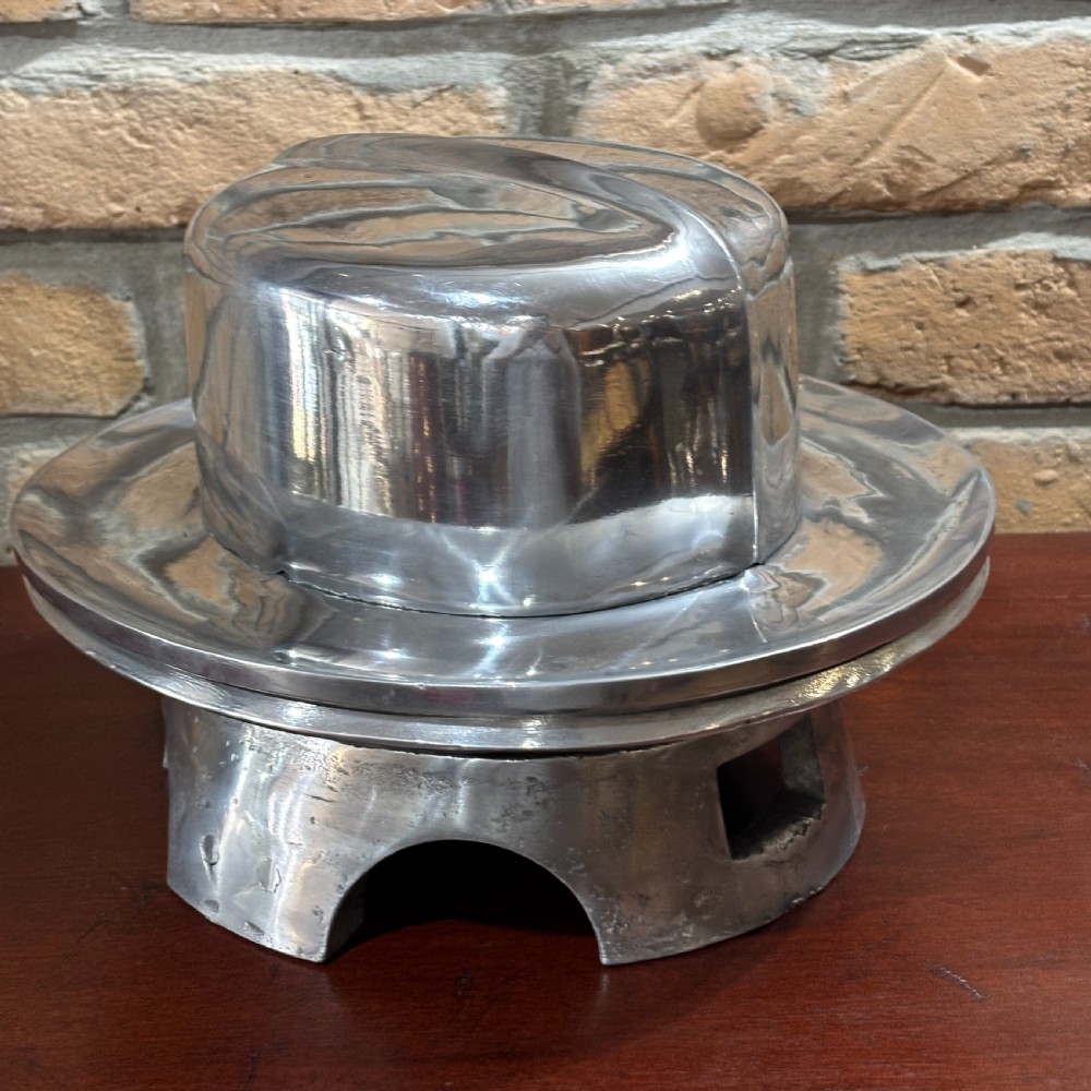 A polished cast aluminium millinery hat block mould forming a fedora shape on a contoured pedestal base. View IV