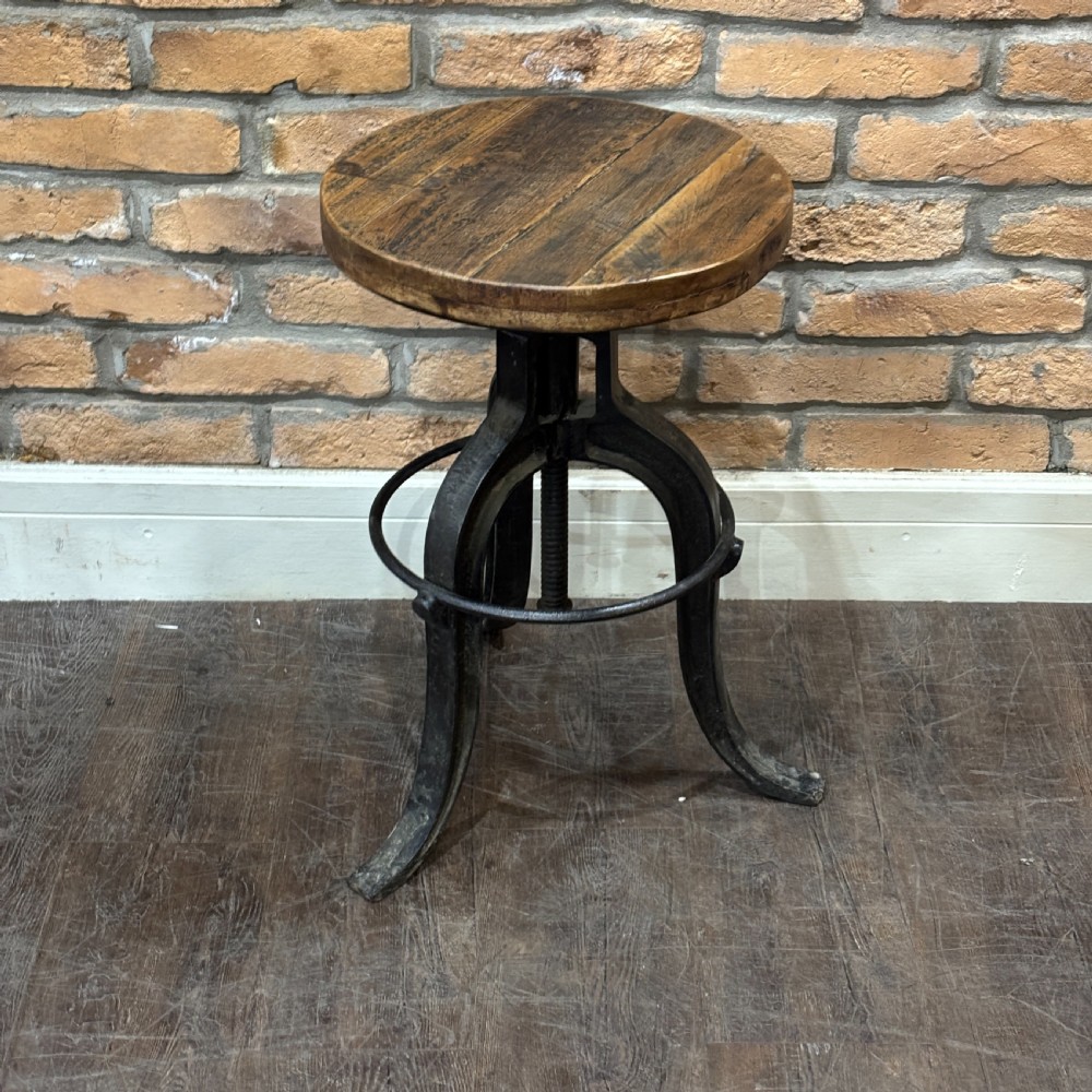 An adjustable industrial workshop stool featuring a circular dark wood seat supported by a black cast iron tripod base with a central screw mechanism and circular footrest.