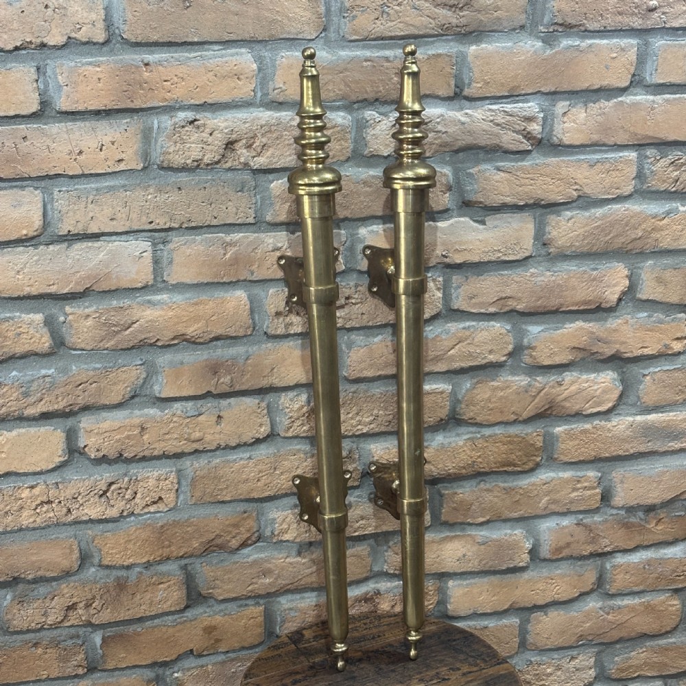 A pair of long, vertical antique brass door pull handles featuring turned urn finials at the top and mounting brackets.