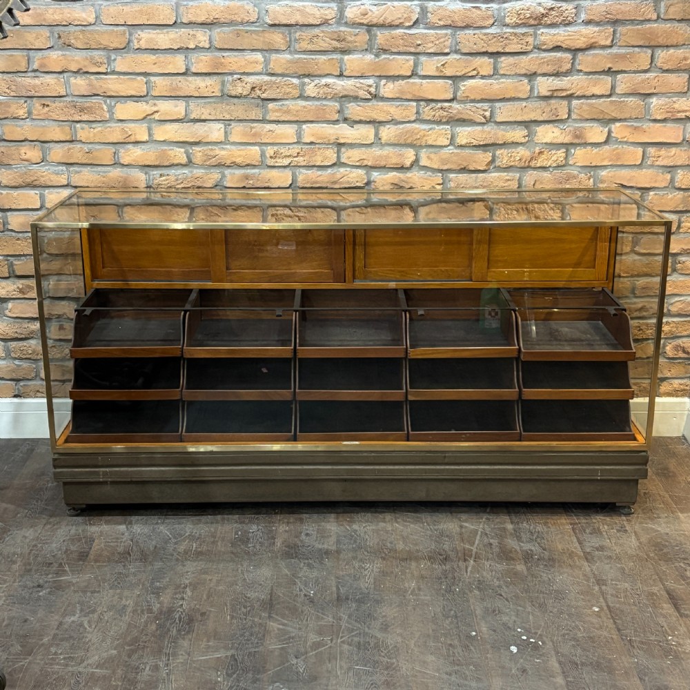 A 1930s brass-framed shop display counter featuring glass side and top panels, tiered interior timber display bins, and timber sliding back doors.
