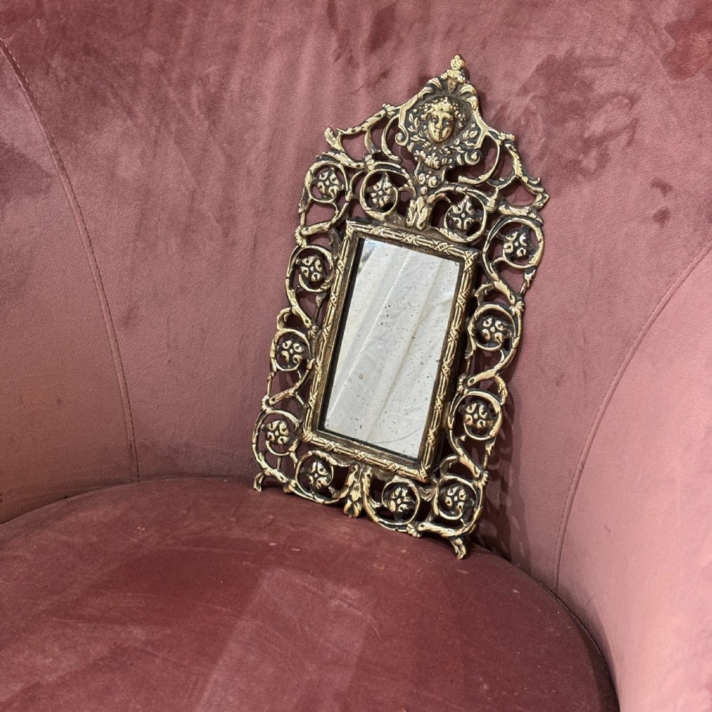 A rectangular glass mirror set within an ornate cast brass frame featuring pierced scrollwork and a central mask crest at the top.