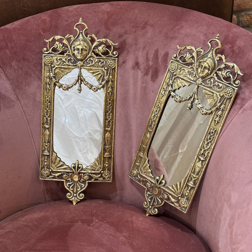 A pair of rectangular brass wall mirrors featuring ornate classical mask crestings, floral garlands, and scrolled detailing.