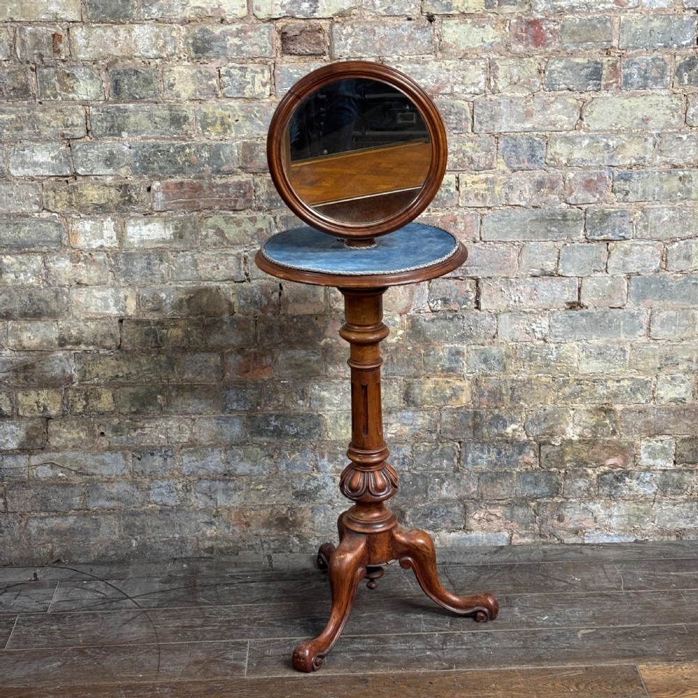 A Victorian mahogany shaving stand featuring a circular tilting mirror mounted above a blue fabric-lined shelf, supported by a turned column and tripod scroll legs.