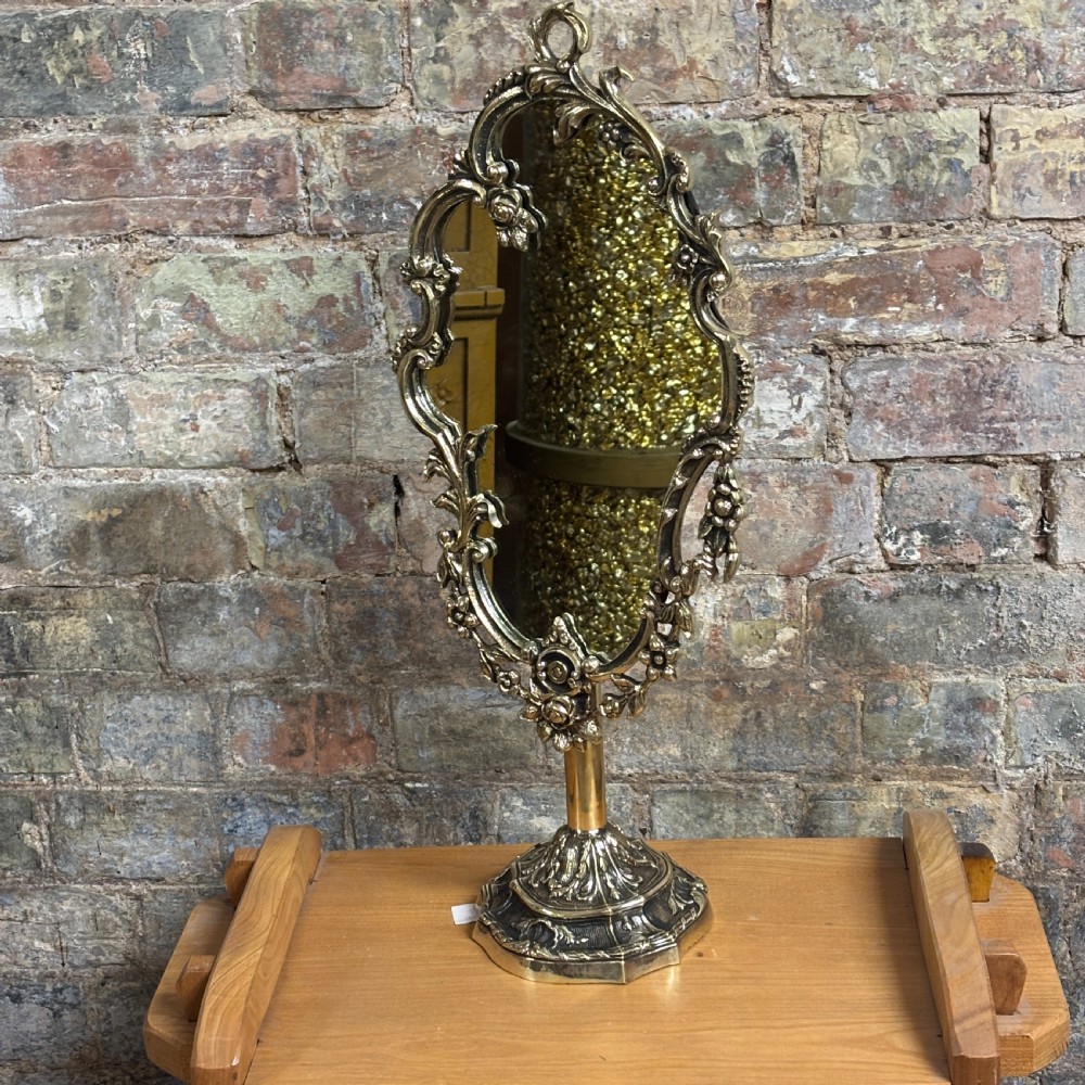 An ornate 1930s cast brass dressing table mirror with detailed floral scrollwork, an oval mirror frame, and a sturdy pedestal base.