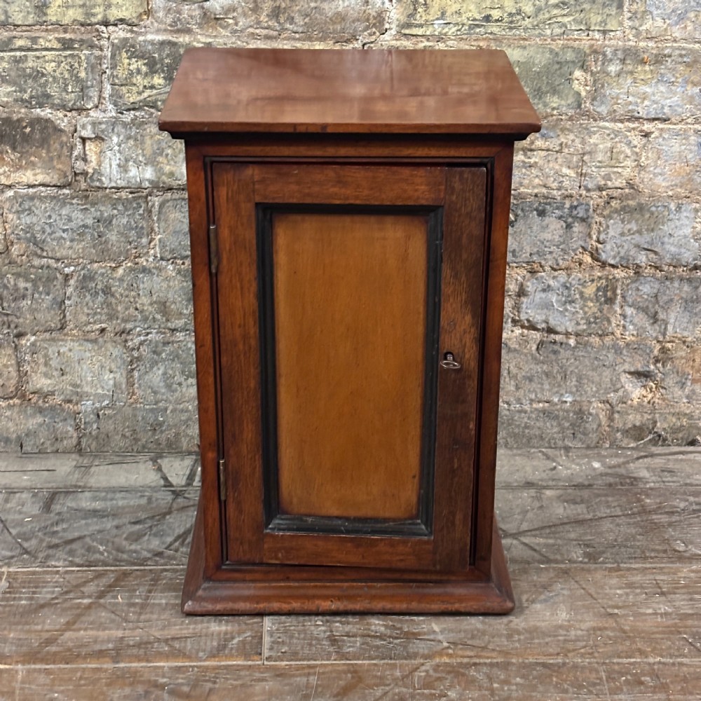 A compact dark mahogany tabletop cabinet featuring a single recessed panel door with a keyhole, a slanted top, and a stepped plinth base.