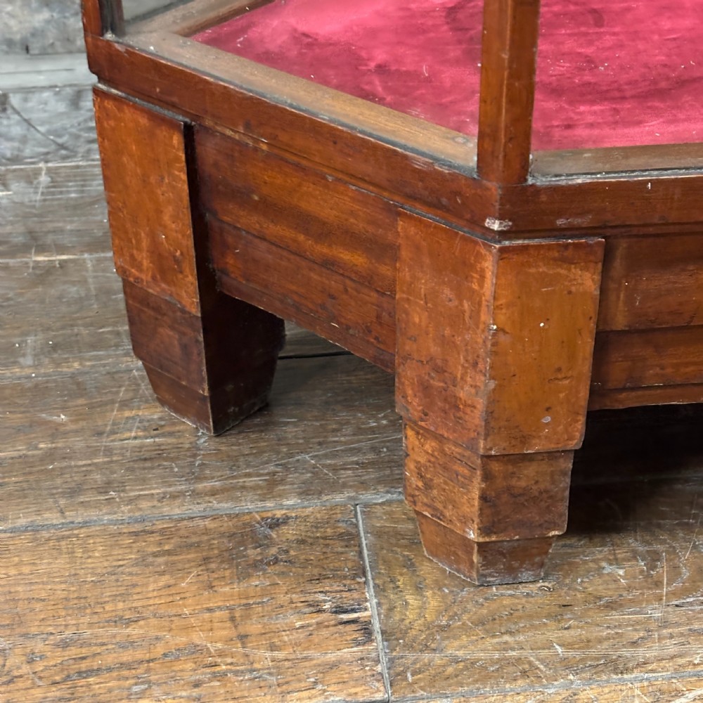 A 1920s pentagonal mahogany display counter featuring glass side panels, a clear glass top, a red velvet-lined interior base, and a central glass shelf. View V