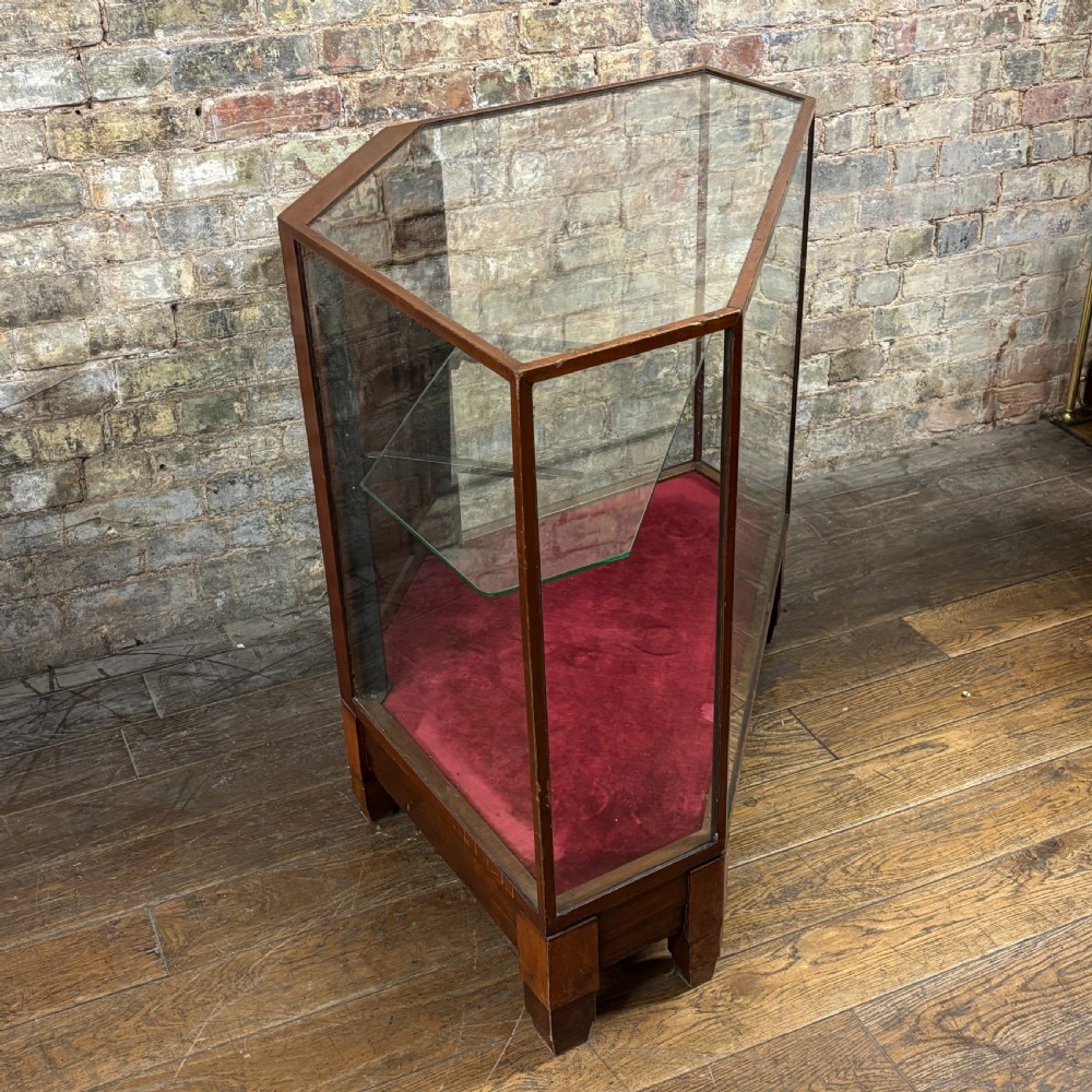 A 1920s pentagonal mahogany display counter featuring glass side panels, a clear glass top, a red velvet-lined interior base, and a central glass shelf. View VI