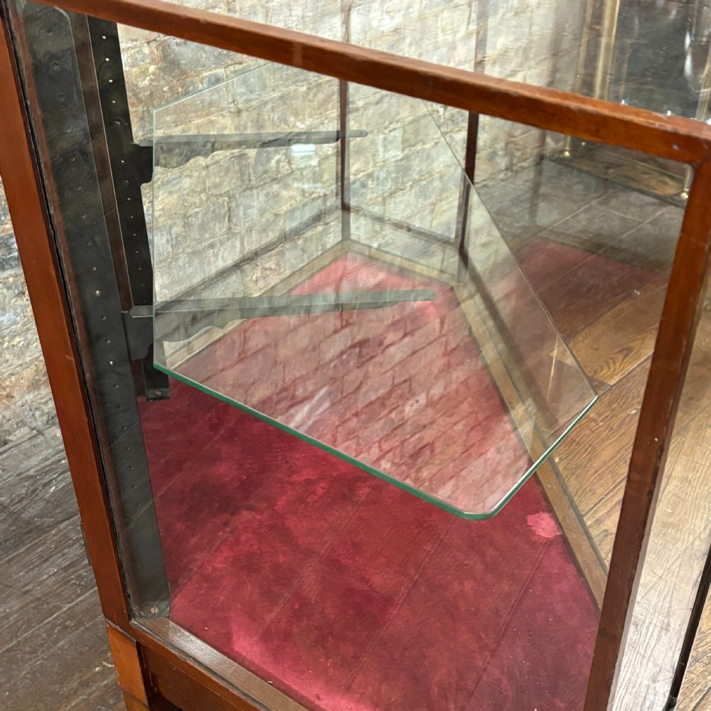A 1920s pentagonal mahogany display counter featuring glass side panels, a clear glass top, a red velvet-lined interior base, and a central glass shelf. View VII