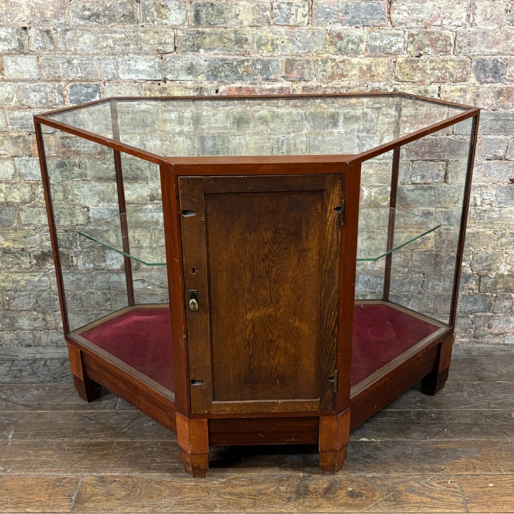 A 1920s pentagonal mahogany display counter featuring glass side panels, a clear glass top, a red velvet-lined interior base, and a central glass shelf. View VIII
