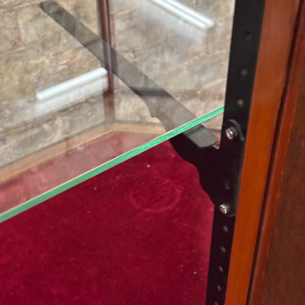 A 1920s pentagonal mahogany display counter featuring glass side panels, a clear glass top, a red velvet-lined interior base, and a central glass shelf. View X