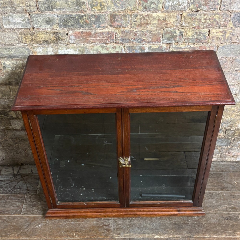 A 1920s dark oak countertop display cabinet featuring twin glazed doors with a central brass catch and internal shelving. View II