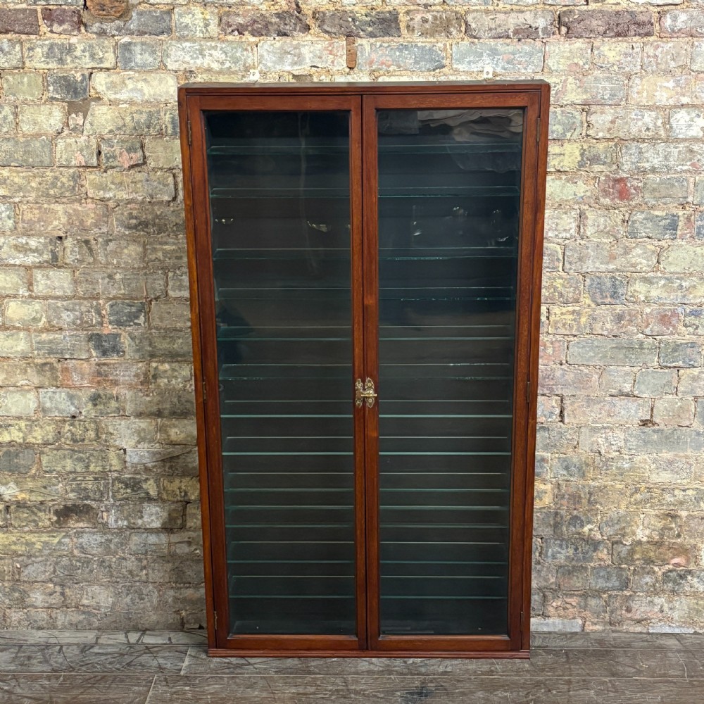 An early 20th-century wall-mounted mahogany cabinet featuring double glazed doors enclosing numerous glass shelves with brass fittings.