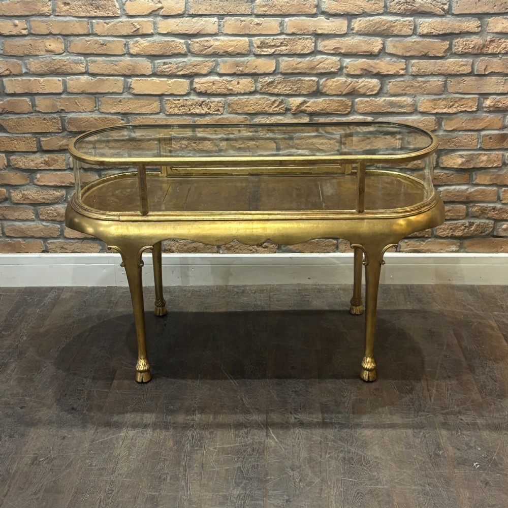 A gilded 1910s oval glass display counter featuring curved side panels and cabriole legs ending in hoof feet.