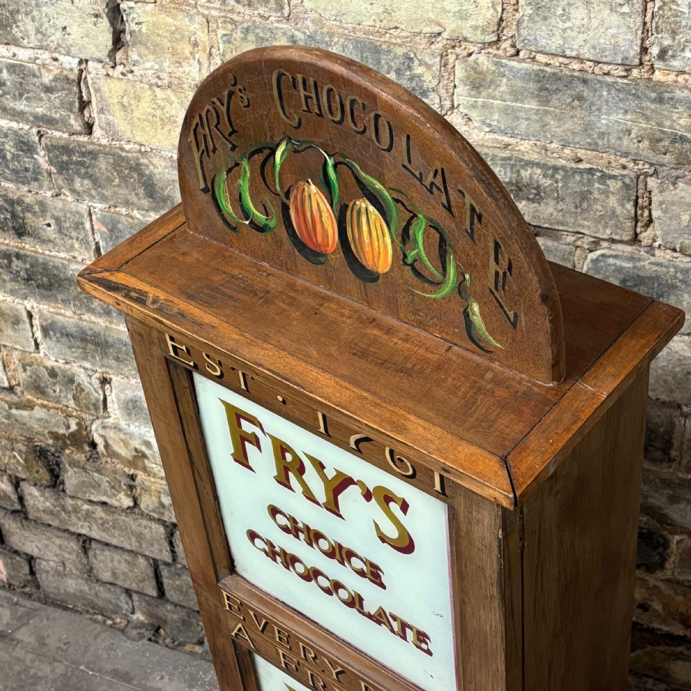 A tall, narrow mahogany advertising display cabinet featuring white glass panels with gold lettering for Frys Chocolate and an arched painted crest. View III