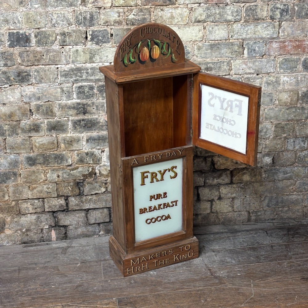 A tall, narrow mahogany advertising display cabinet featuring white glass panels with gold lettering for Frys Chocolate and an arched painted crest. View VII