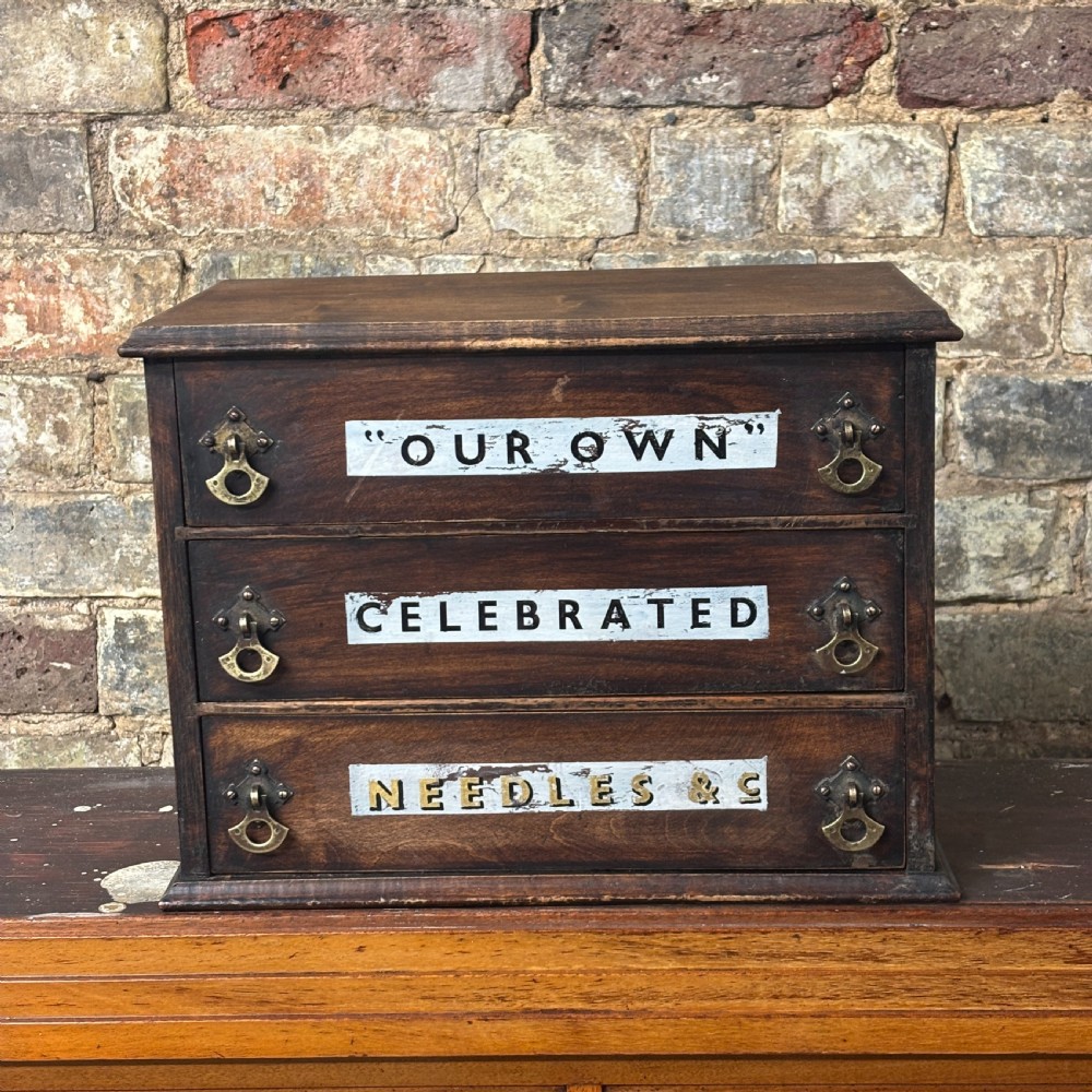 A dark oak three-drawer tabletop drapers cabinet featuring hand-painted commercial typography and brass drop handles.