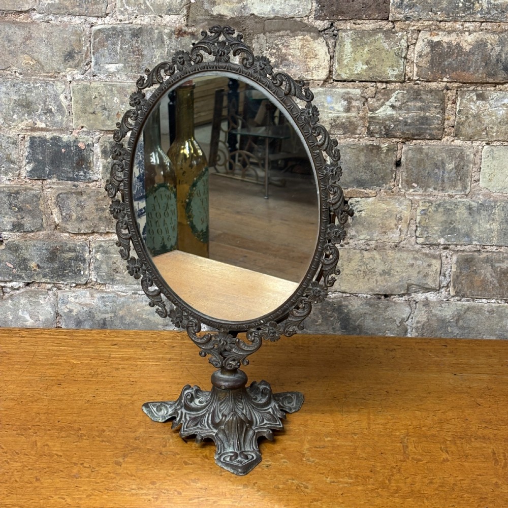 A late Victorian cast bronze dressing table mirror featuring an oval glass frame adorned with intricate floral scrollwork set upon an ornate pedestal base.