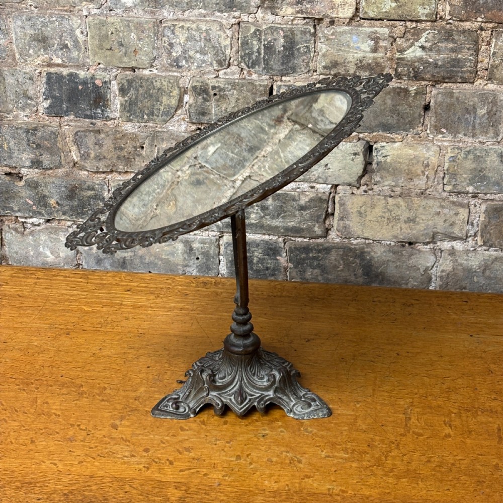 A late Victorian cast bronze dressing table mirror featuring an oval glass frame adorned with intricate floral scrollwork set upon an ornate pedestal base. View II