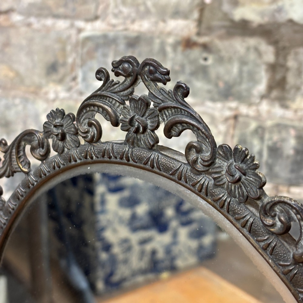 A late Victorian cast bronze dressing table mirror featuring an oval glass frame adorned with intricate floral scrollwork set upon an ornate pedestal base. View IV