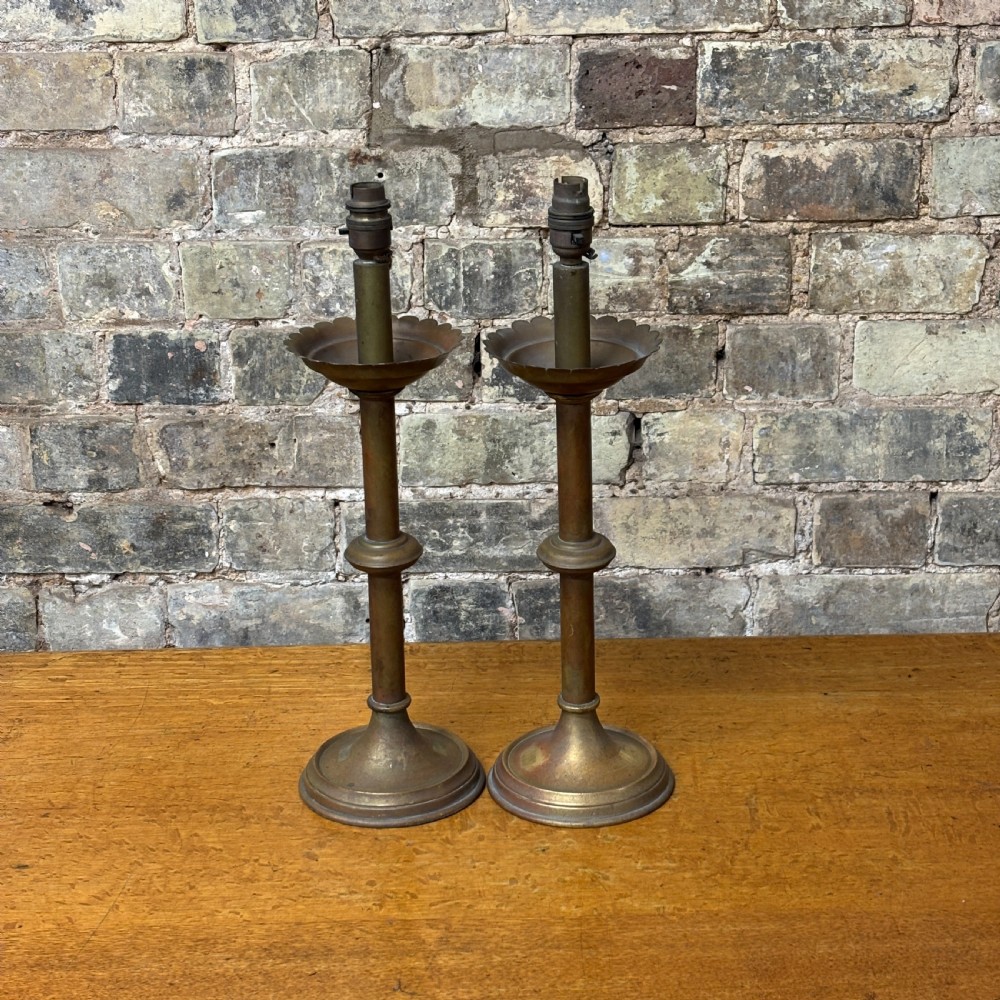 A pair of early 20th-century brass candlestick table lamps with scalloped drip trays, turned stems, and circular stepped bases.