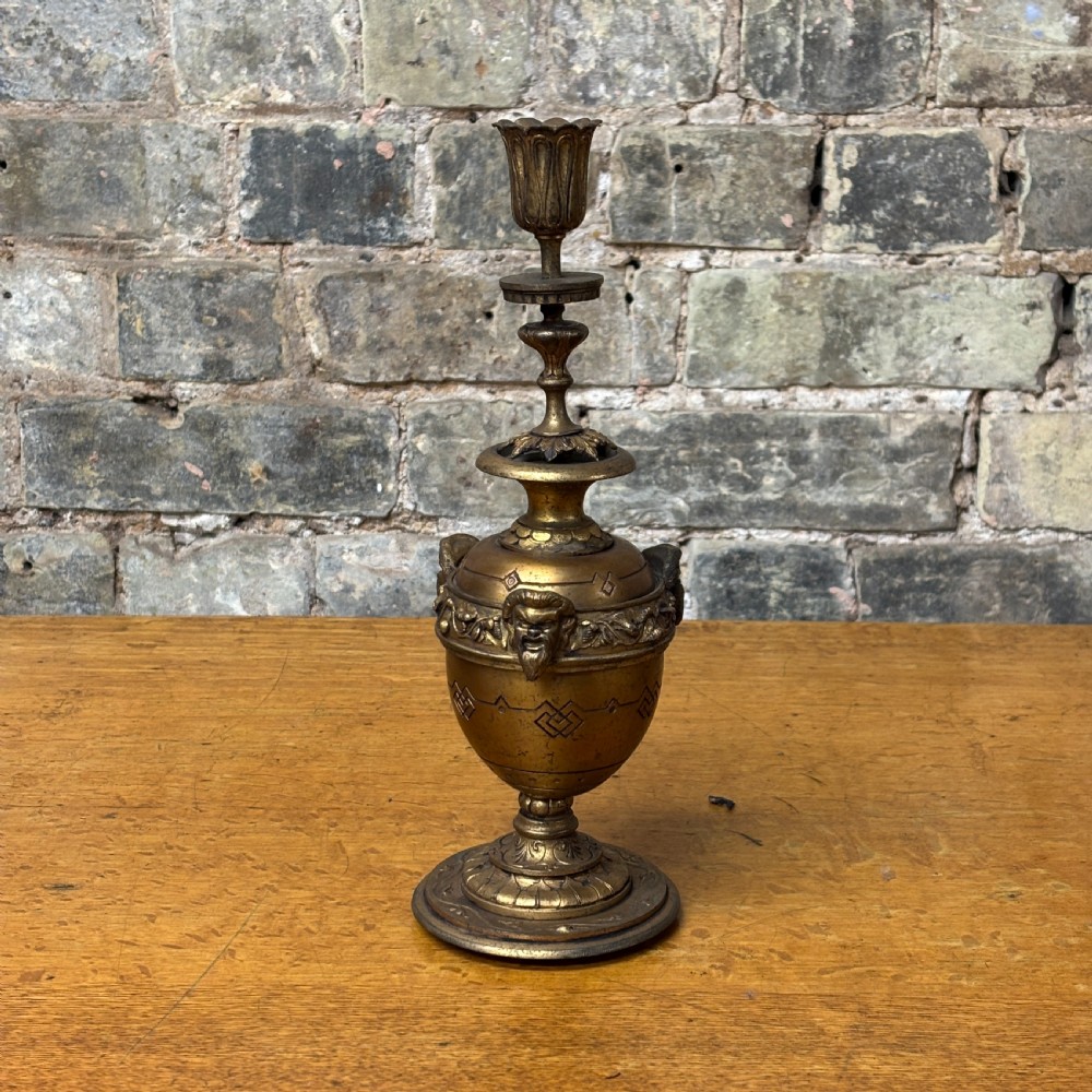 An antique cast brass candlestick featuring an urn-shaped central section with relief mask details, a stepped turned stem, and a fluted nozzle.