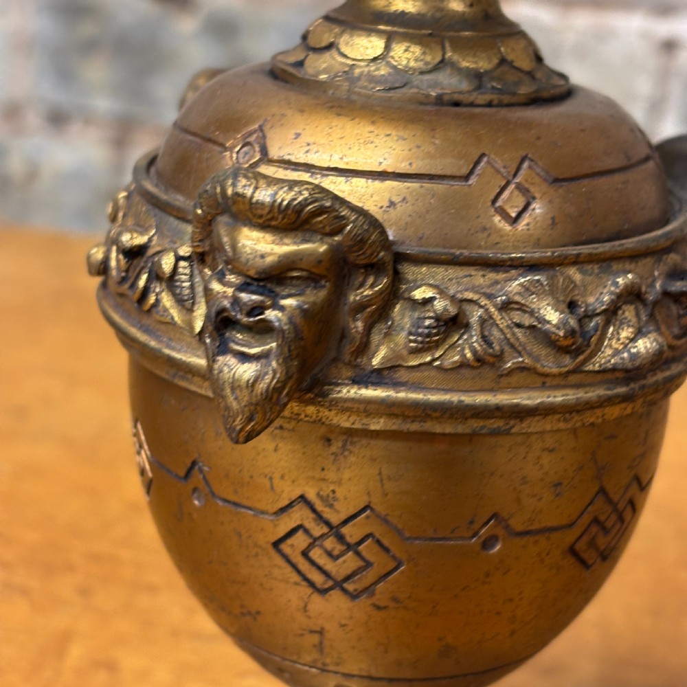 An antique cast brass candlestick featuring an urn-shaped central section with relief mask details, a stepped turned stem, and a fluted nozzle. View III