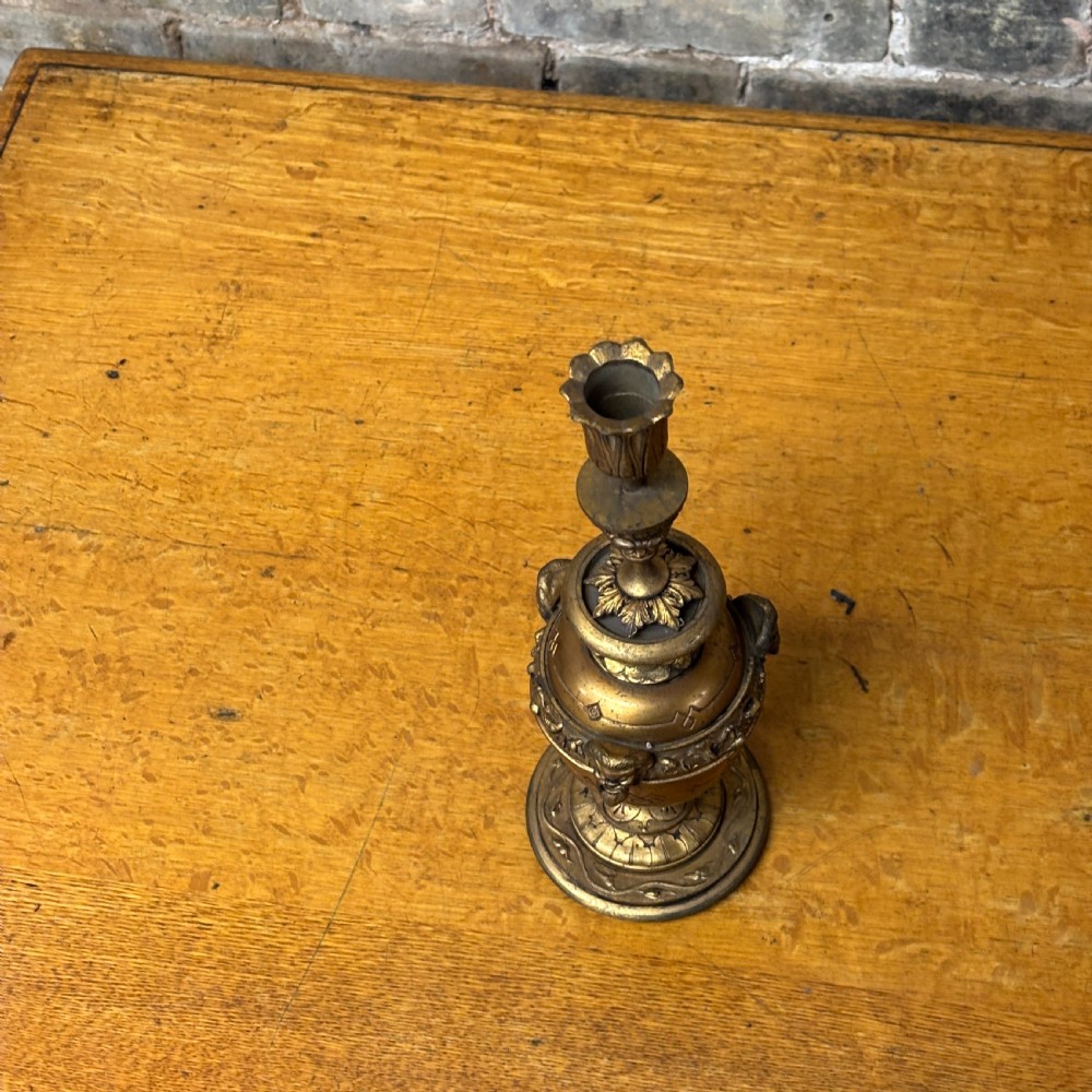 An antique cast brass candlestick featuring an urn-shaped central section with relief mask details, a stepped turned stem, and a fluted nozzle. View V