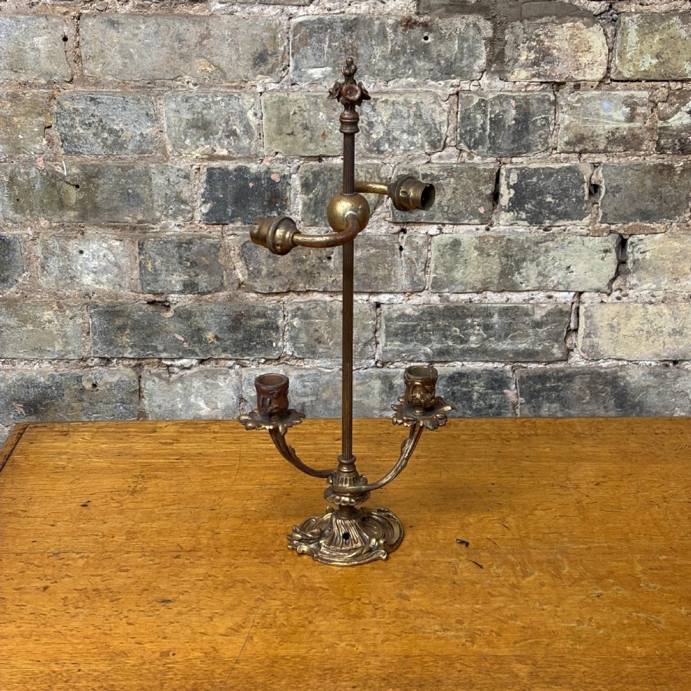 An antique cast brass candelabra table lamp featuring a scrolled base, twin candle sconces, two electrical fittings, and a finial top.