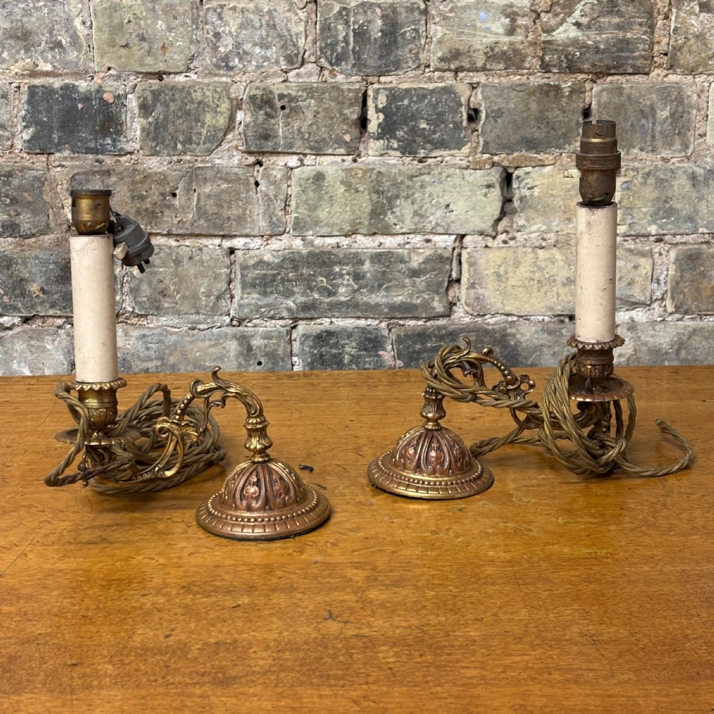 A pair of early 20th-century cast brass wall sconces featuring ornate scrollwork arms, gadrooned backplates, and cream-coloured candle sleeves.