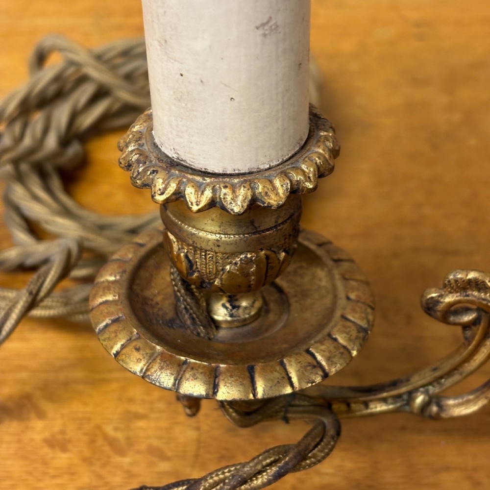 A pair of early 20th-century cast brass wall sconces featuring ornate scrollwork arms, gadrooned backplates, and cream-coloured candle sleeves. View IV