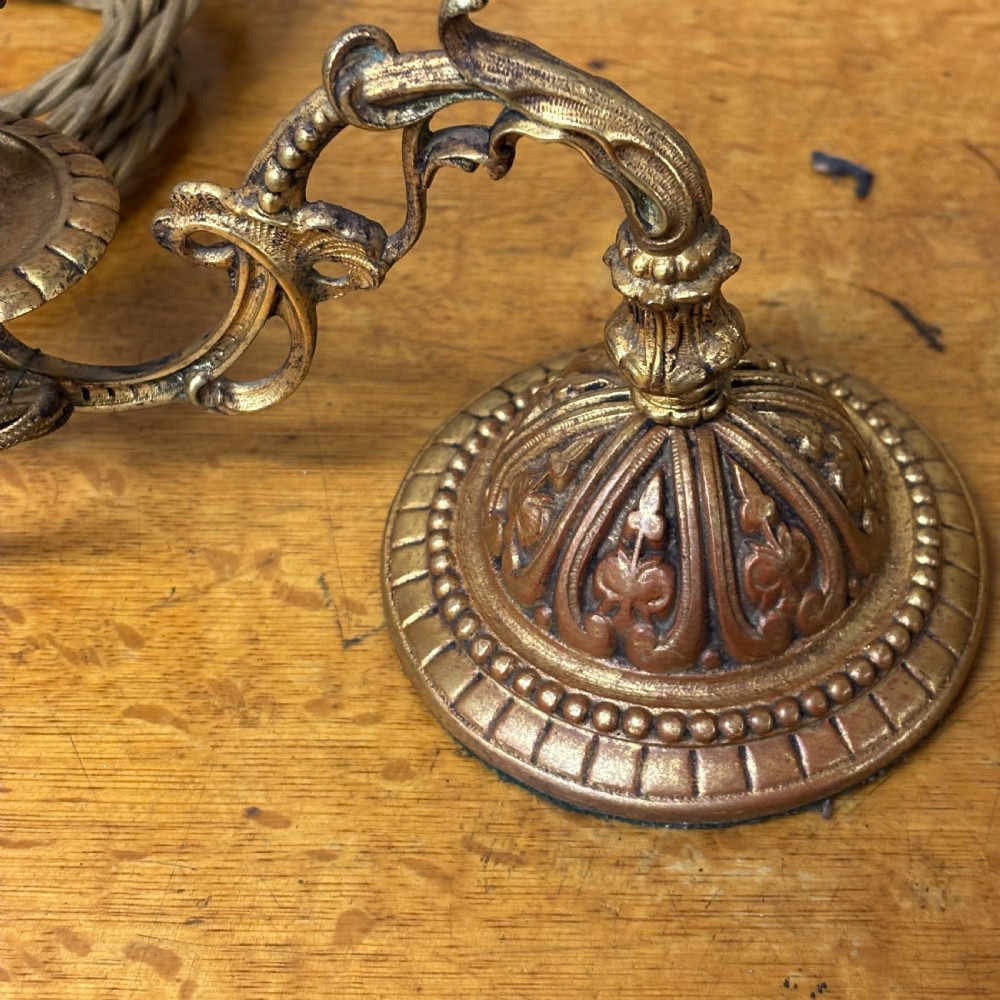 A pair of early 20th-century cast brass wall sconces featuring ornate scrollwork arms, gadrooned backplates, and cream-coloured candle sleeves. View V