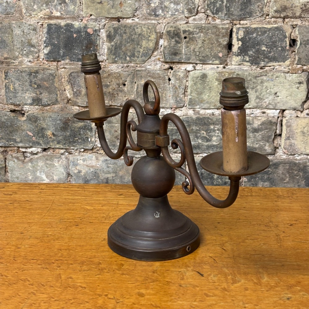 An early 20th-century patinated brass two-arm candelabra table lamp featuring curved scroll arms, faux candle sleeves, and a stepped circular base.