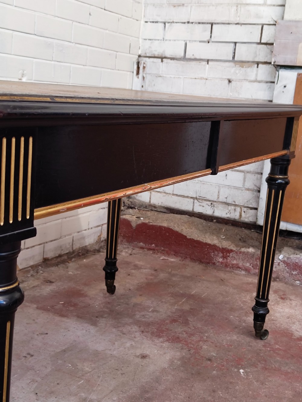 A Victorian Aesthetic Movement ebonised writing table featuring a brown leather inset top, fluted legs with gilt detailing, and brass castors. View II