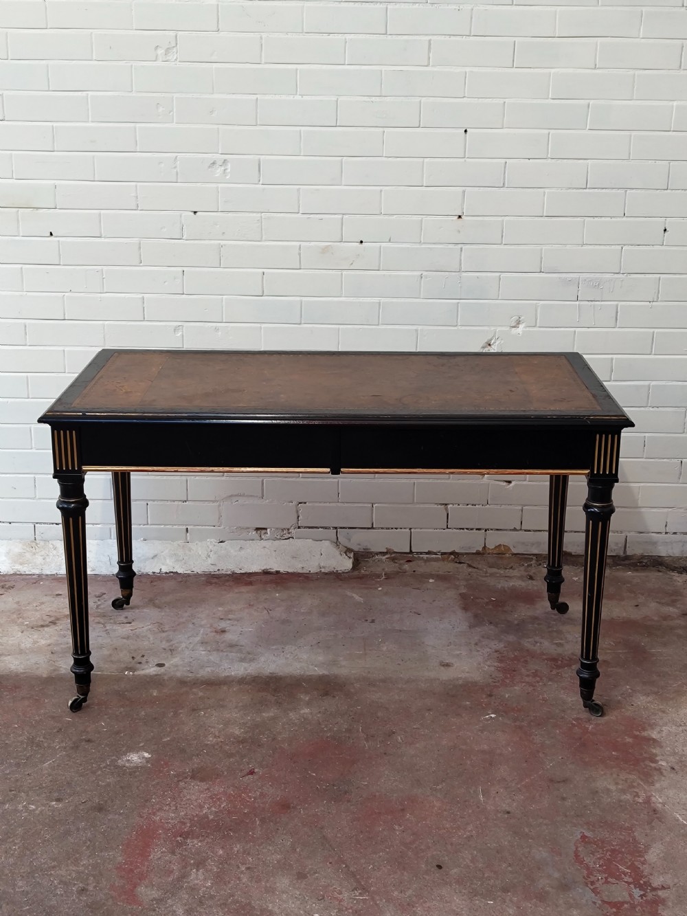 A Victorian Aesthetic Movement ebonised writing table featuring a brown leather inset top, fluted legs with gilt detailing, and brass castors. View IV