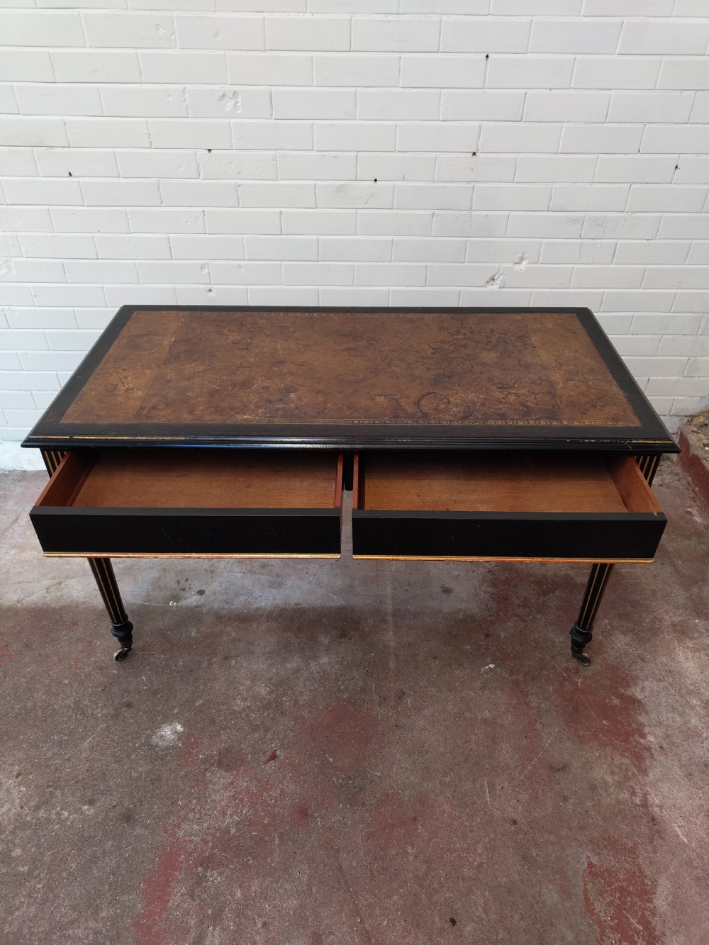 A Victorian Aesthetic Movement ebonised writing table featuring a brown leather inset top, fluted legs with gilt detailing, and brass castors. View V