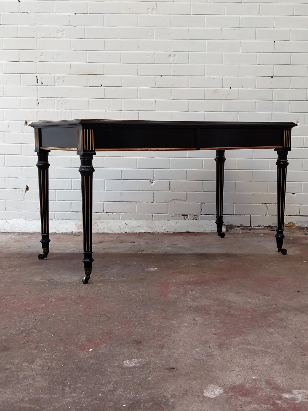 A Victorian Aesthetic Movement ebonised writing table featuring a brown leather inset top, fluted legs with gilt detailing, and brass castors. View XIV