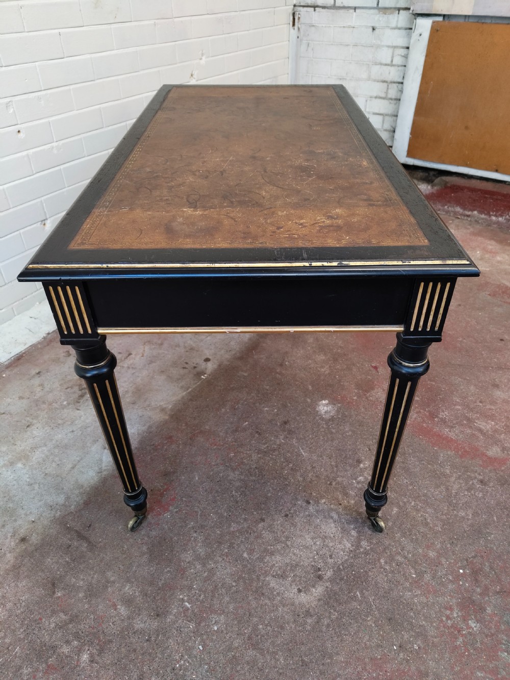 A Victorian Aesthetic Movement ebonised writing table featuring a brown leather inset top, fluted legs with gilt detailing, and brass castors. View XVI