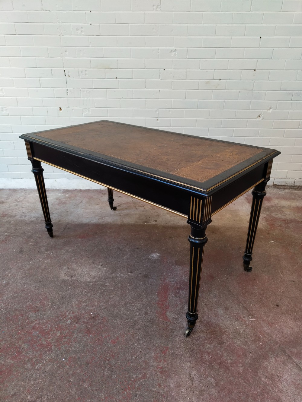 A Victorian Aesthetic Movement ebonised writing table featuring a brown leather inset top, fluted legs with gilt detailing, and brass castors. View XVII
