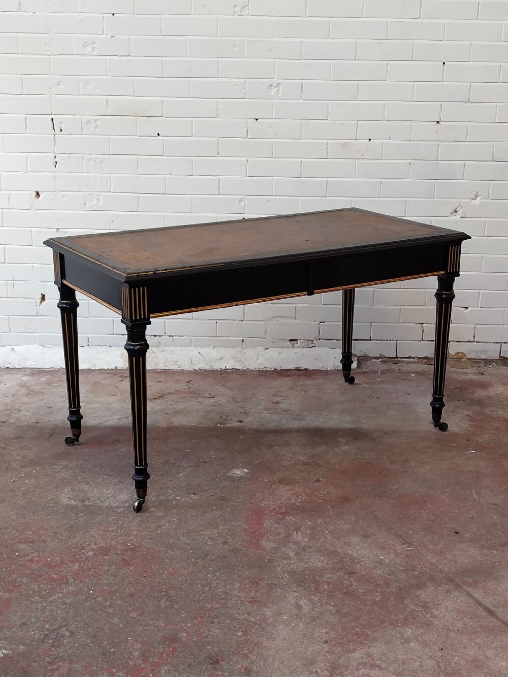 A Victorian Aesthetic Movement ebonised writing table featuring a brown leather inset top, fluted legs with gilt detailing, and brass castors. View XIX