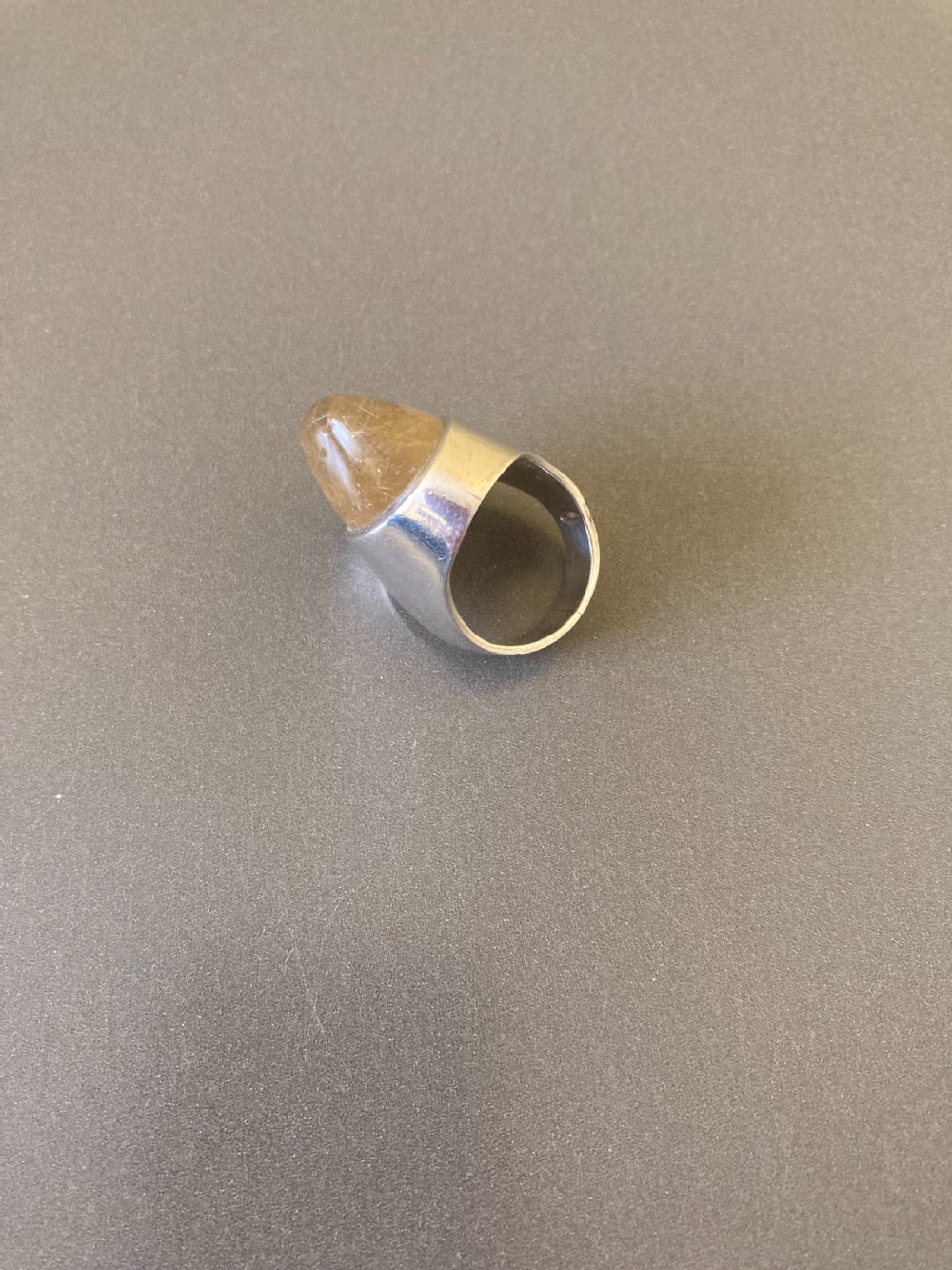 A close-up shot of a vintage Danish silver ring featuring a smooth, domed rutilated quartz gemstone set in a heavy sterling silver band, resting on a small clear acrylic display stand. View V