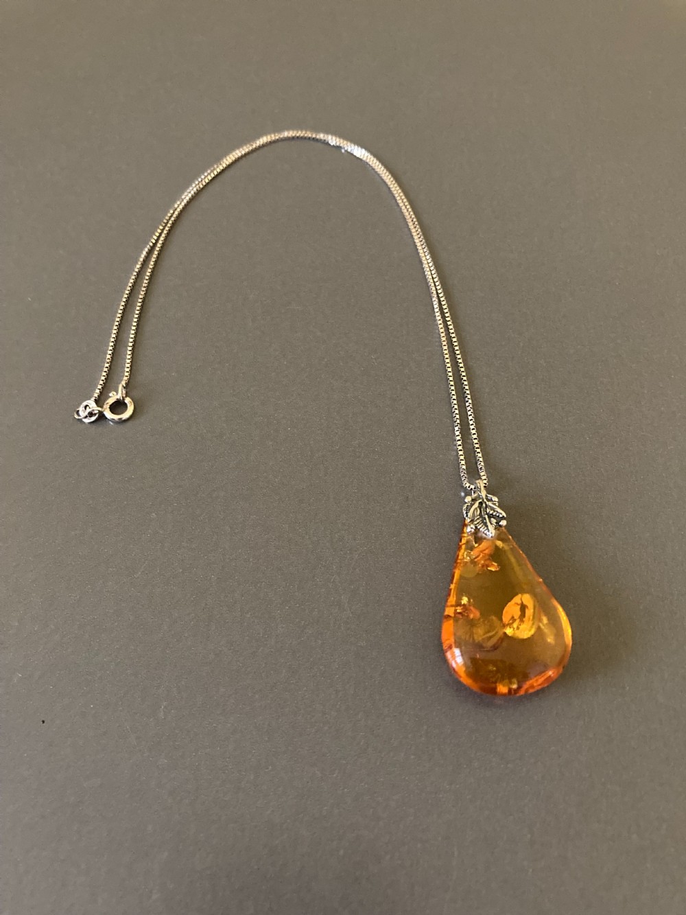 A teardrop-shaped amber pendant hangs from a thin silver chain against a neutral grey surface.