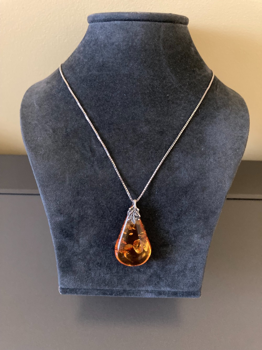 A teardrop-shaped amber pendant hangs from a thin silver chain against a neutral grey surface. View III